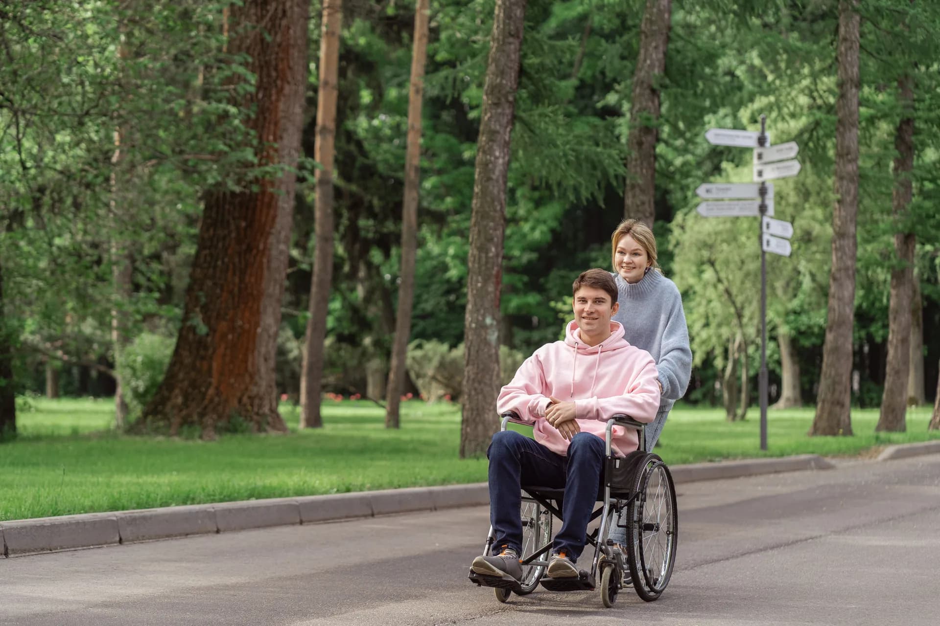 A woman assisting a young man using a wheelchair along a park path